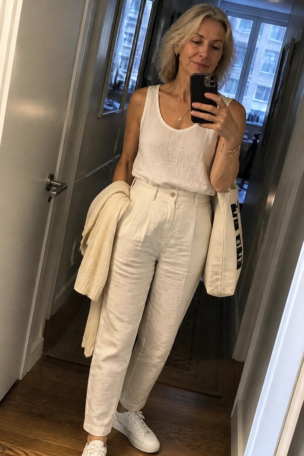 Woman with gray hair in white linen outfit holding phone, sweater, and tote bag