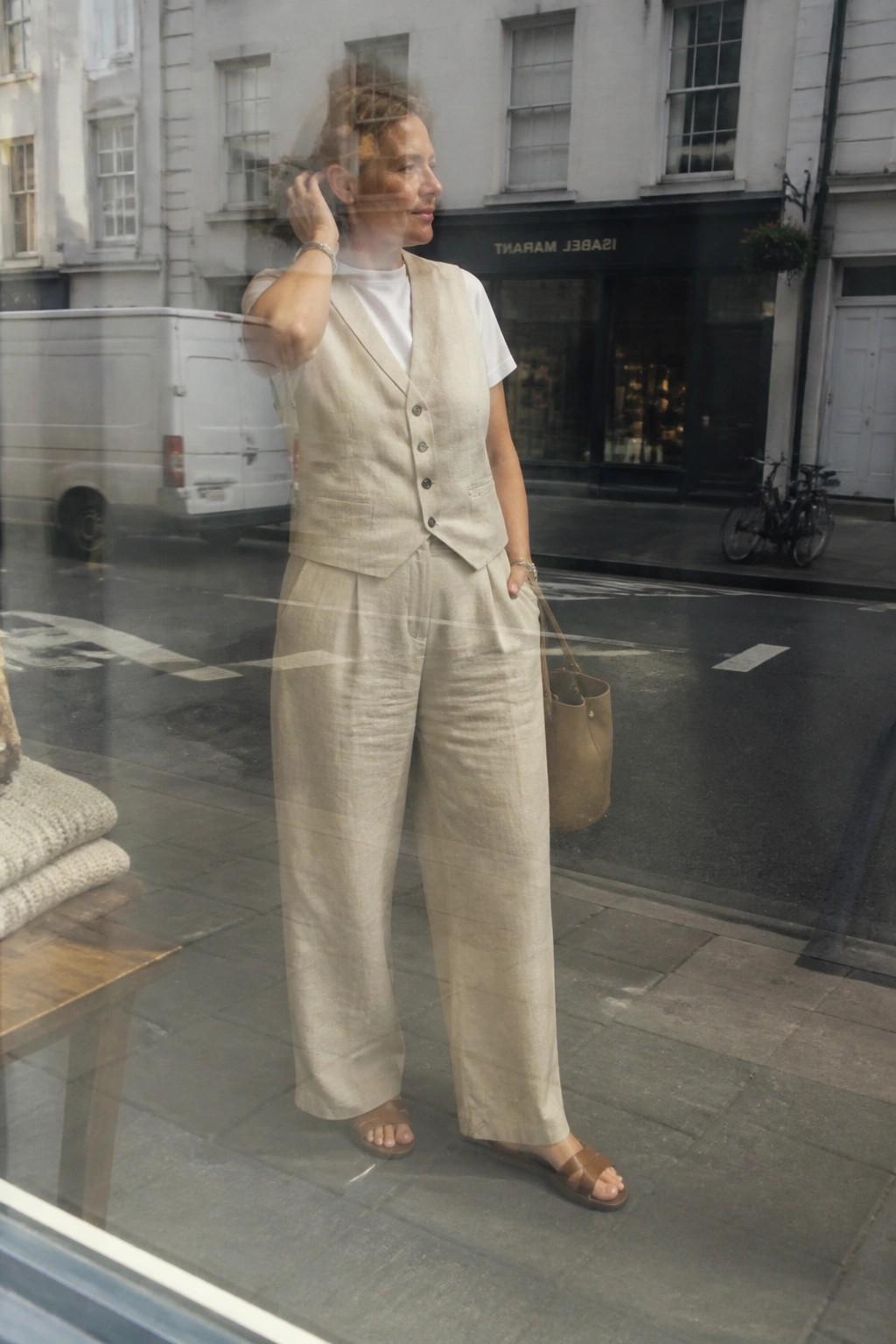 Woman in beige linen vest and trousers touches hair on city street