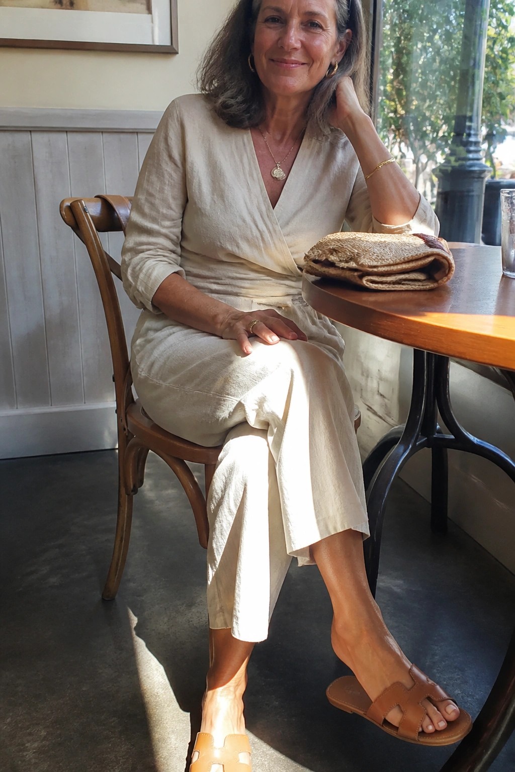 Woman in cream linen wrap outfit at cafe table with woven clutch and leather sandals