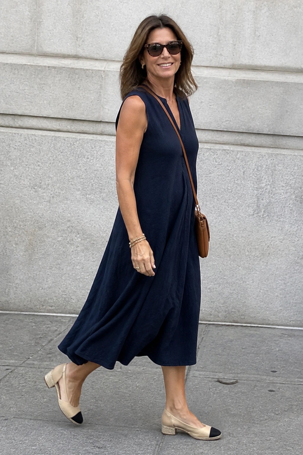 Woman wearing a navy sleeveless linen midi dress with V neckline, brown leather crossbody bag, beige cap toe block heel flats, layered bracelets, and sunglasses while walking on pavement.