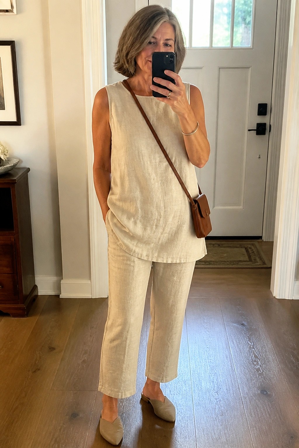 Woman wearing a sleeveless cream linen tank top and matching straight-leg pants with a brown crossbody bag and beige mules, taking a mirror selfie indoors.