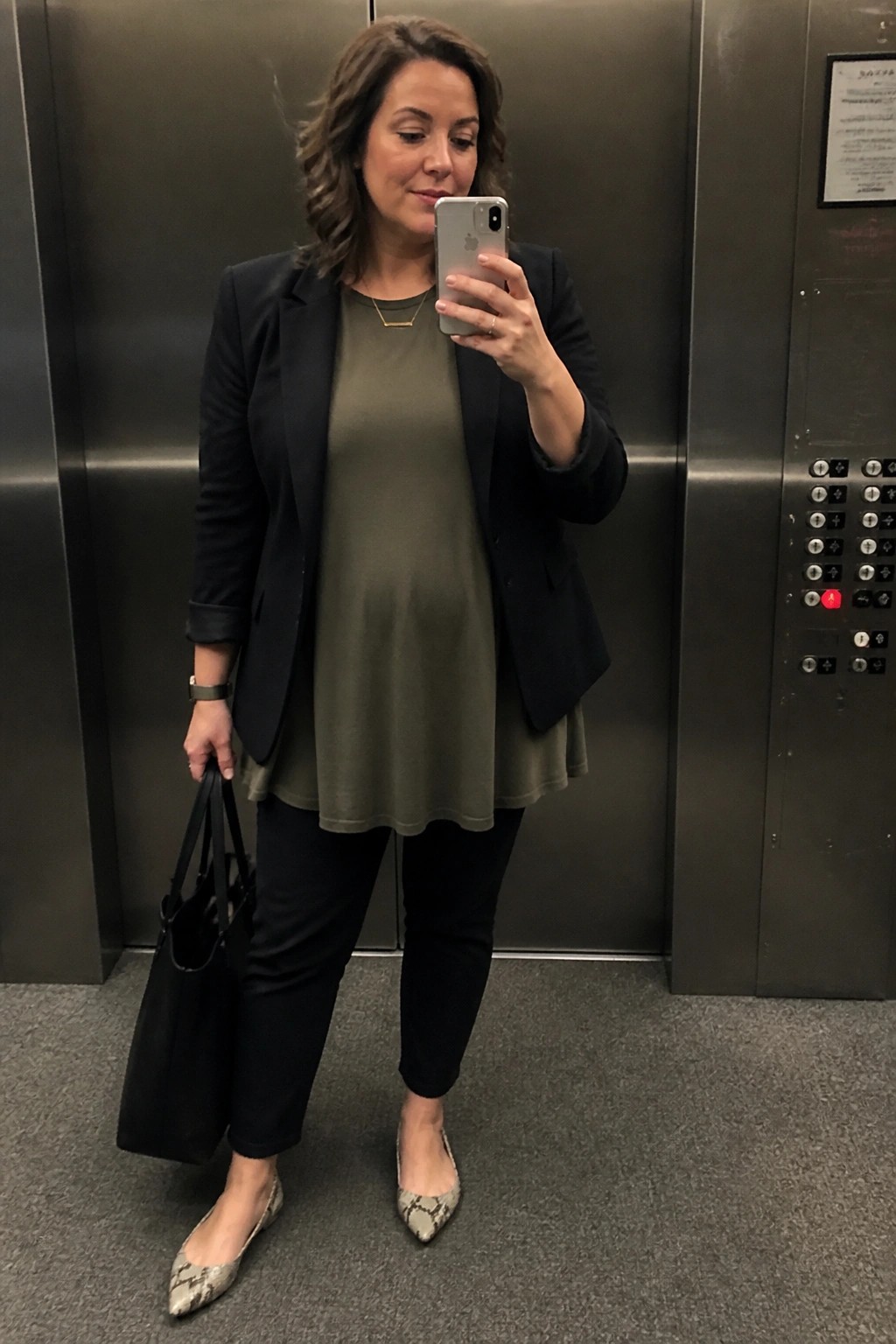 Woman in black blazer and olive top taking selfie in elevator with tote bag