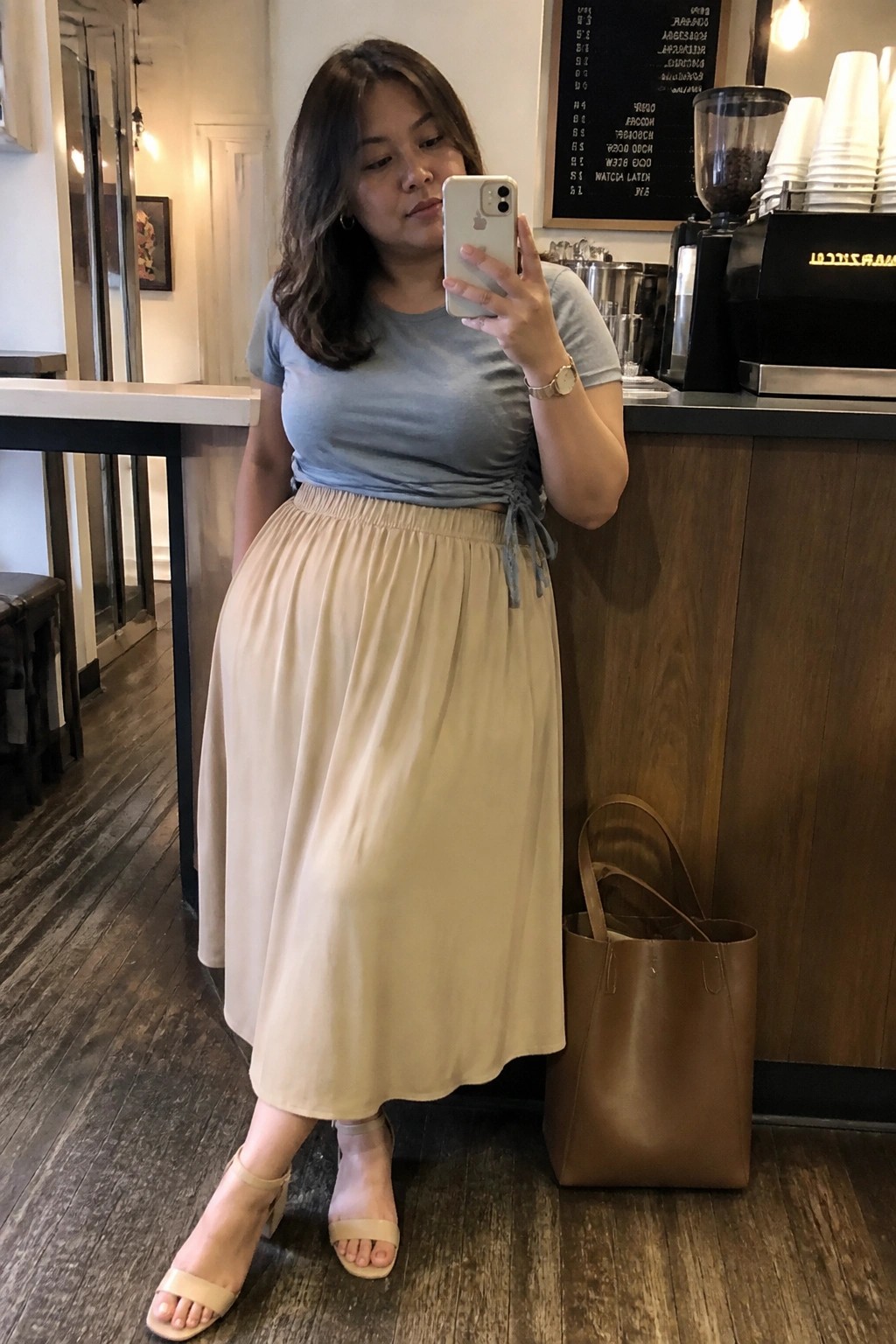 Woman in light blue tied t-shirt and flowing beige midi skirt with block heel sandals holding phone for mirror selfie