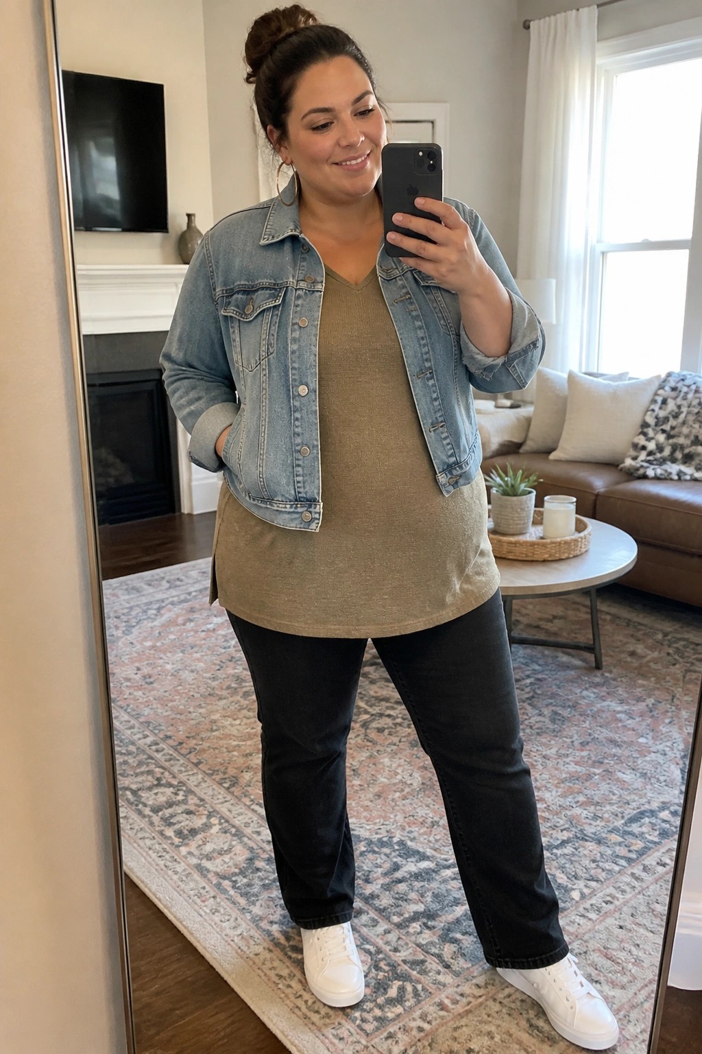 Plus size woman in a light blue denim jacket over an olive green long tunic top paired with black straight leg pants and white sneakers taking a mirror selfie indoors.