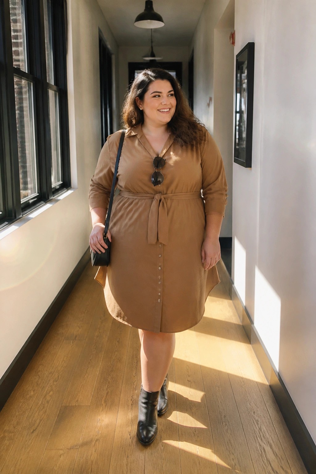 Smiling woman in brown belted shirt dress and black boots in sunlit hallway.