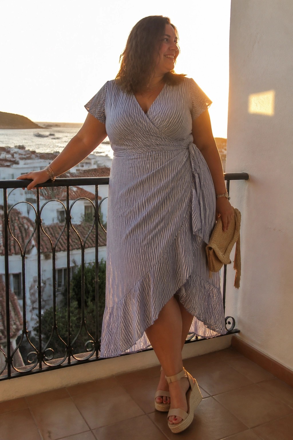 Woman in striped wrap dress on balcony overlooking sea at sunset