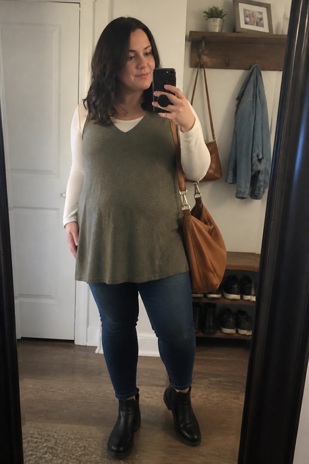 Woman taking mirror selfie in olive green top, jeans, and black boots