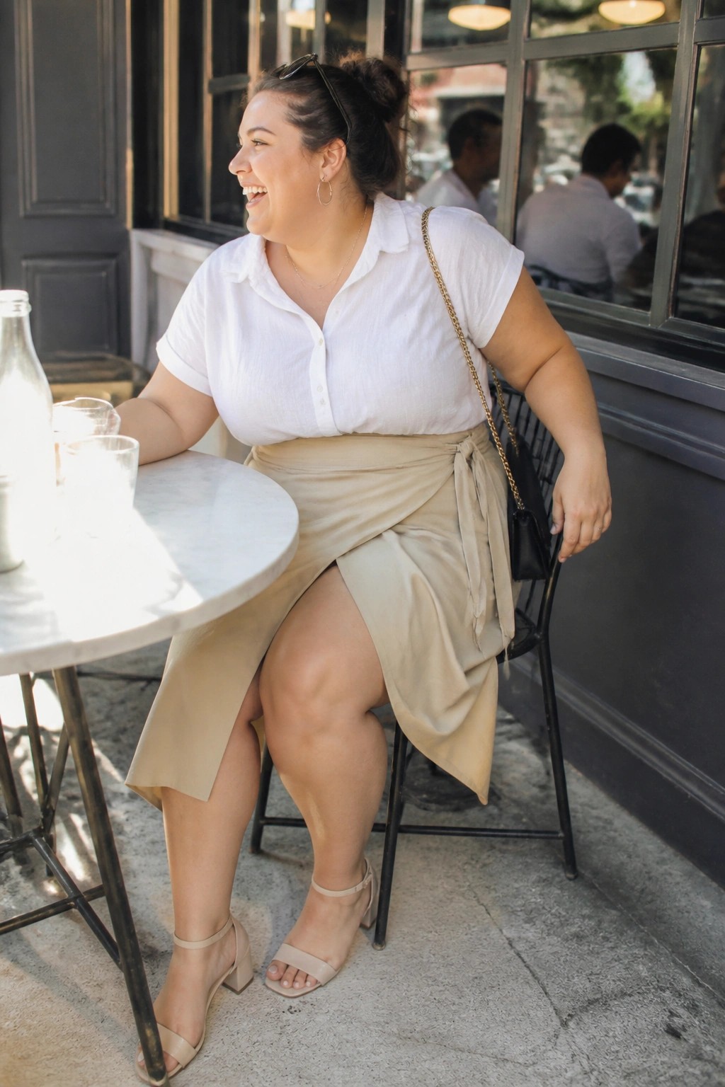 Plus size woman wearing a white short sleeve button up shirt tucked into a beige wrap midi skirt with side tie, paired with matching block heel sandals and a black chain strap purse.