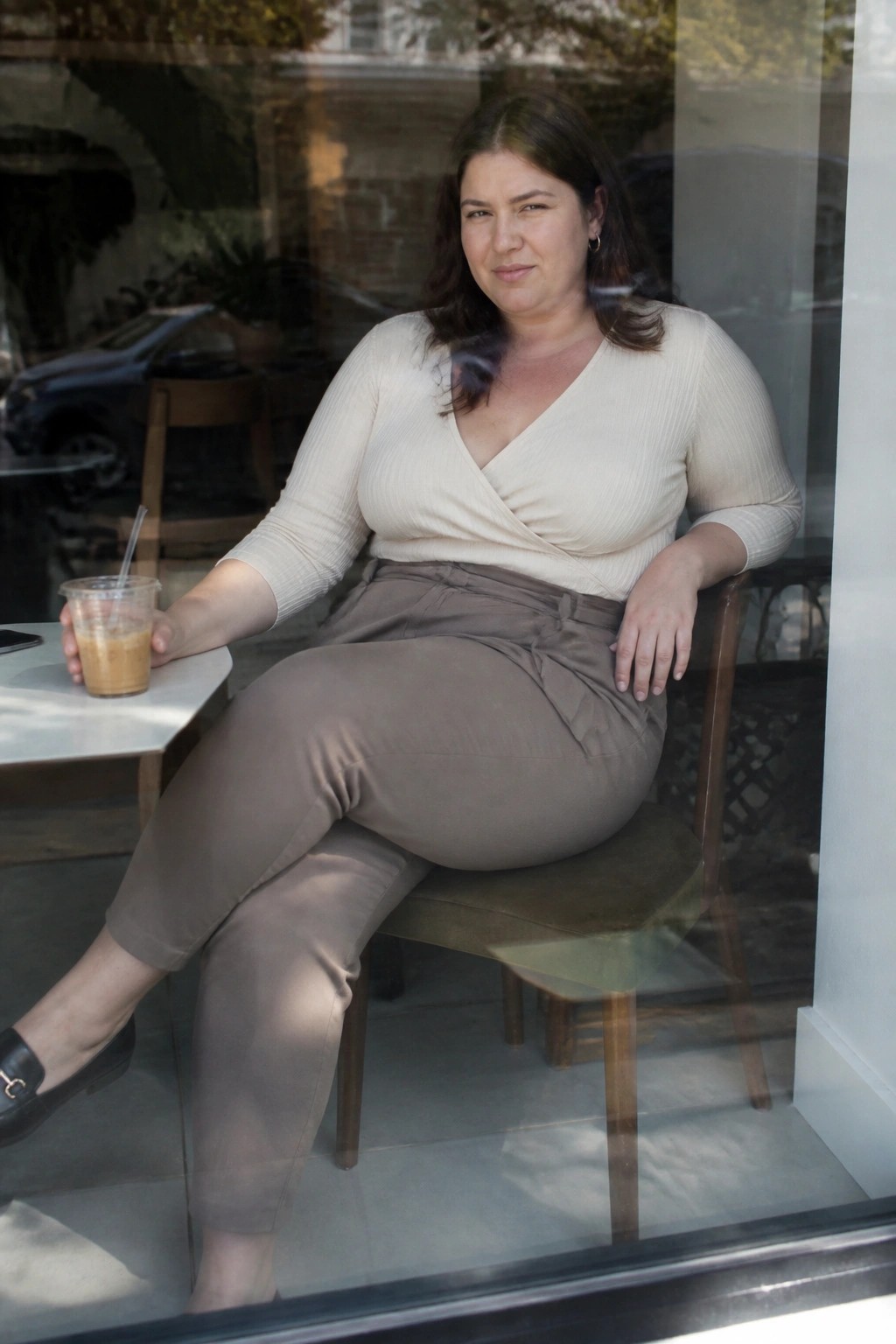 Woman in beige wrap top and brown pants sits by window holding iced coffee