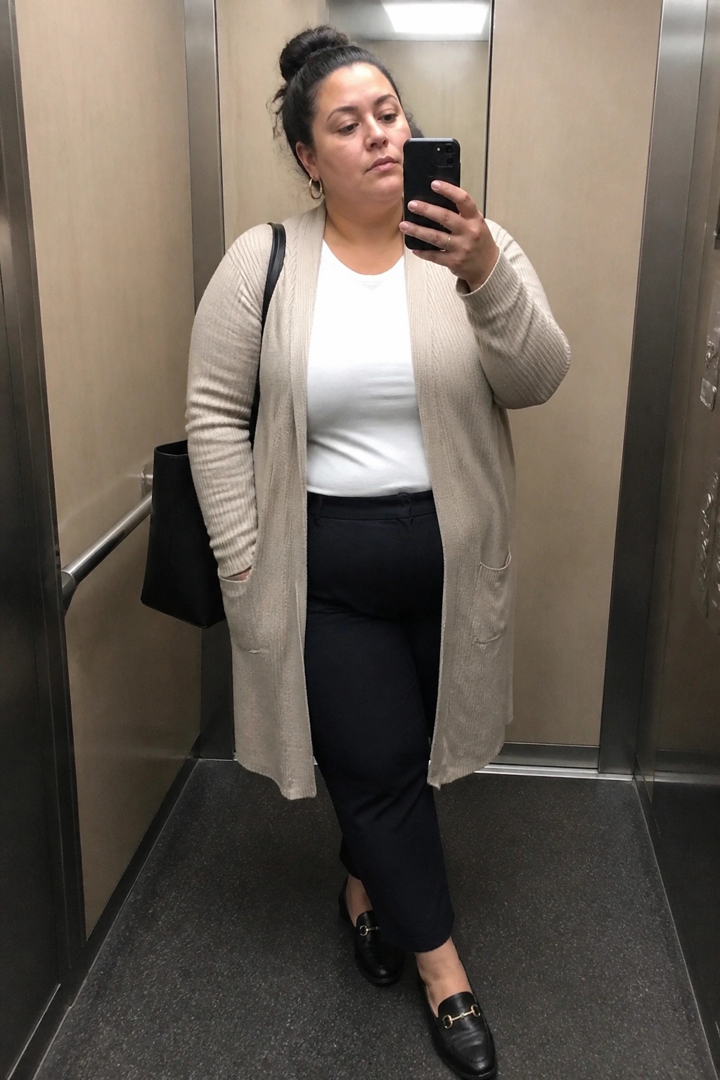 Woman in beige cardigan and black pants taking mirror selfie in elevator