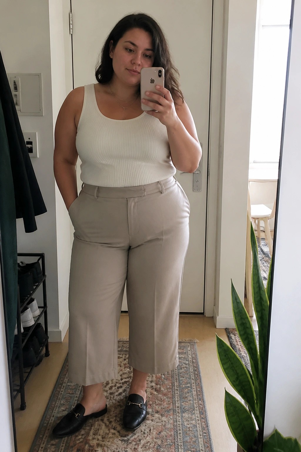 Woman in white tank top and beige pants taking mirror selfie with phone.