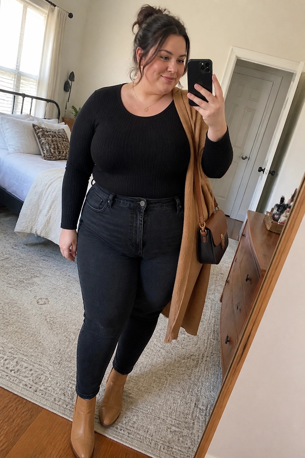 Plus size woman wearing a black ribbed long sleeve top tucked into dark wash jeans with a tan open cardigan draped over her shoulders, tan ankle boots, and a brown crossbody bag while taking a mirror selfie in a bedroom.