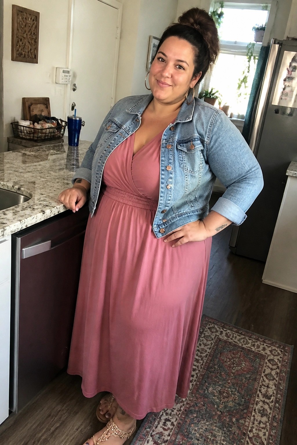Woman in pink dress and denim jacket stands in kitchen leaning on counter