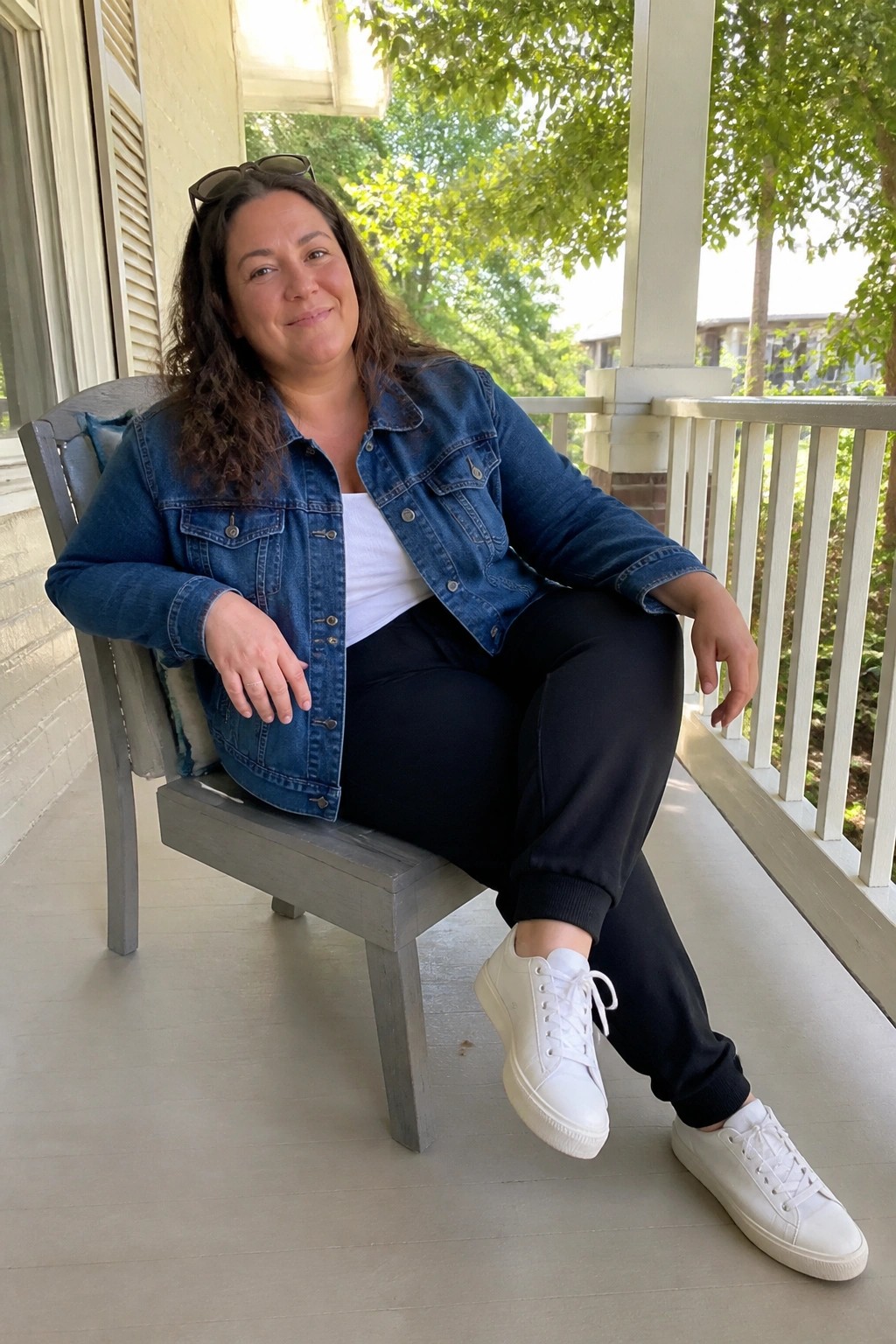 Woman in blue denim jacket and white sneakers sits on porch in black pants