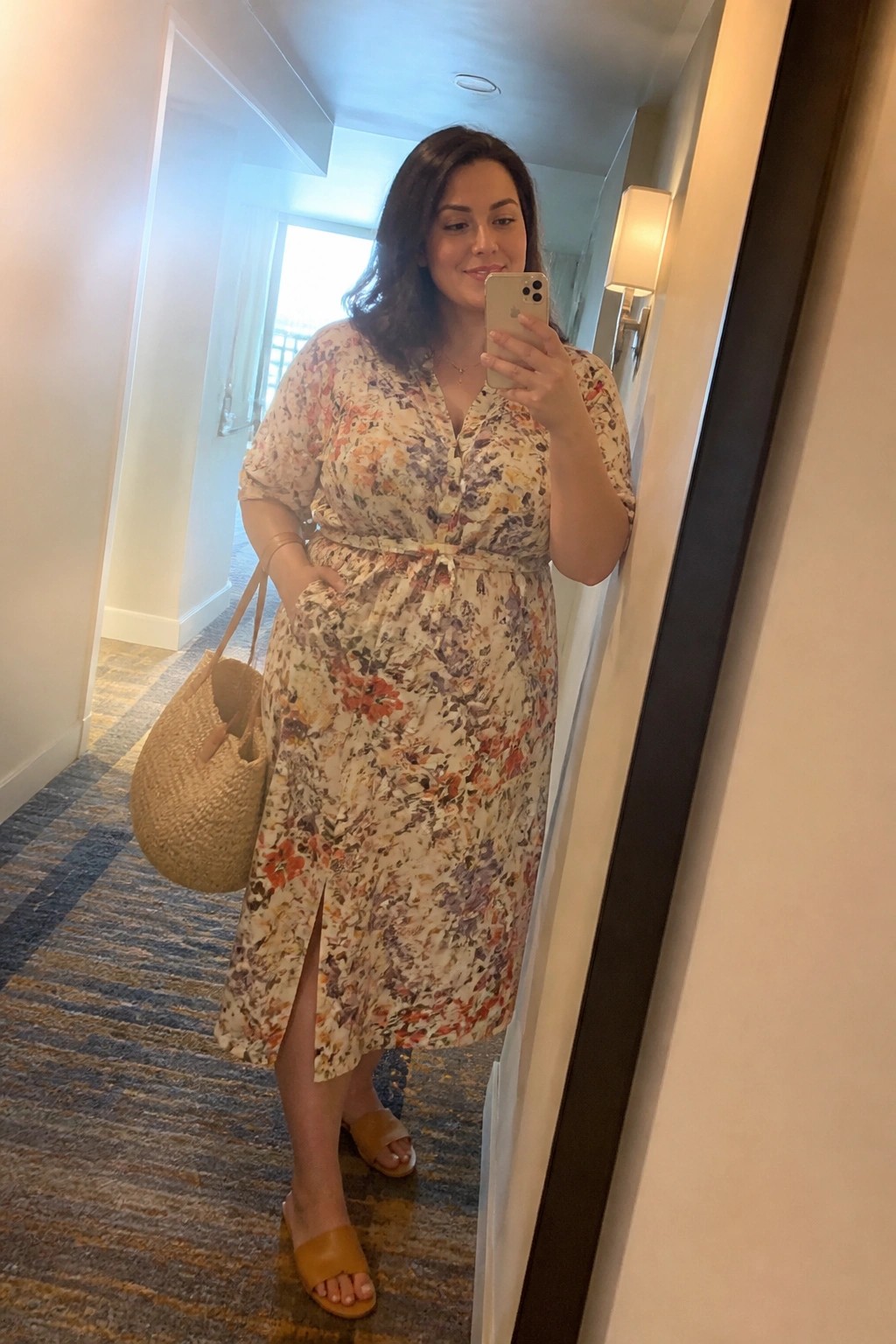 Woman in floral midi dress taking mirror selfie with woven bag in hallway
