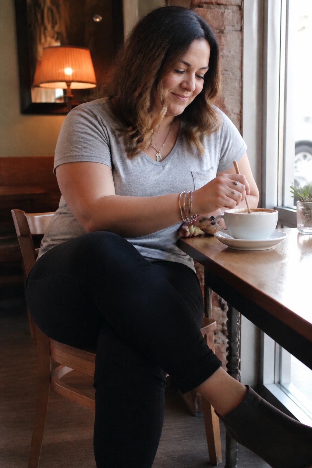 Woman in gray t-shirt stirring coffee at wooden cafe table by window