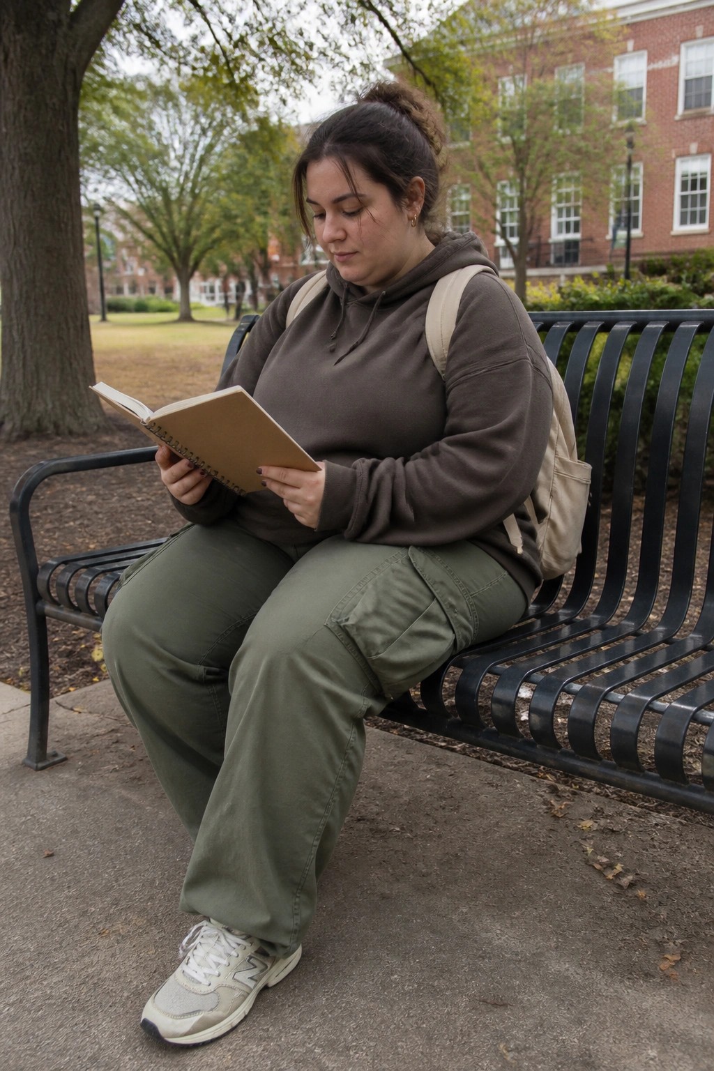 Woman seated on a bench wearing an oversized brown hoodie, olive green cargo pants, beige backpack, and light gray sneakers while holding an open notebook.