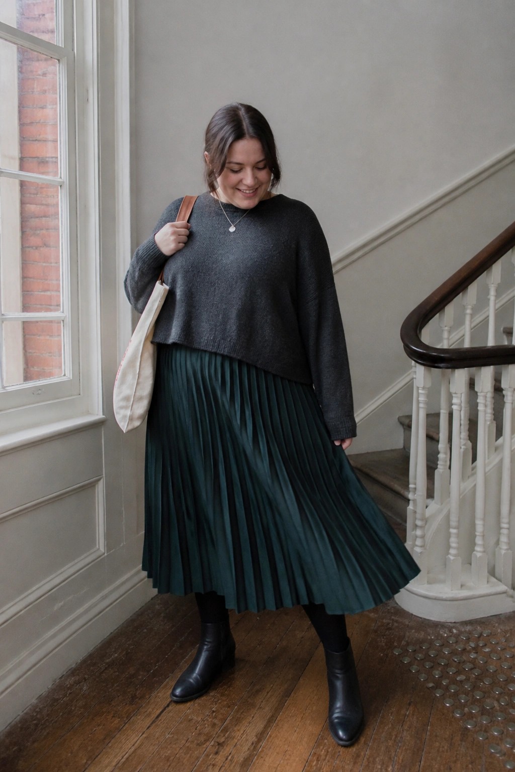 Woman in gray sweater and green pleated skirt with tote bag by staircase