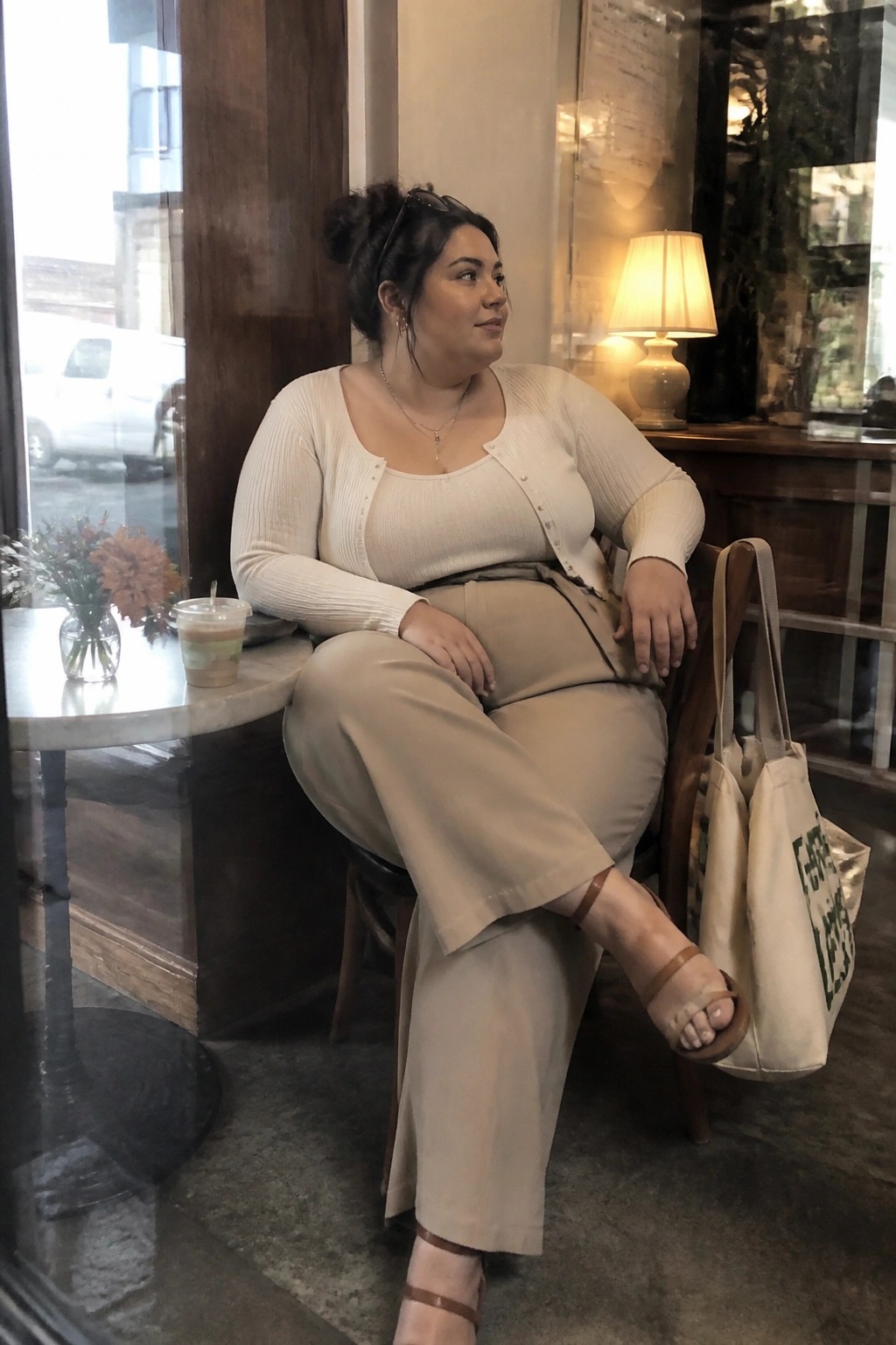 Woman in cream cardigan and beige pants seated in cafe with tote bag.