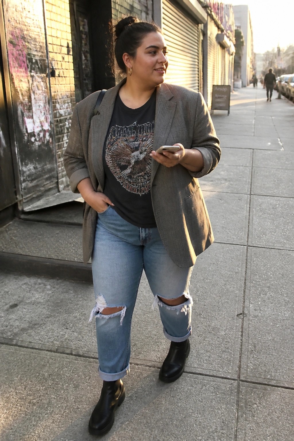 Woman in gray blazer and ripped jeans holds phone on sunny sidewalk