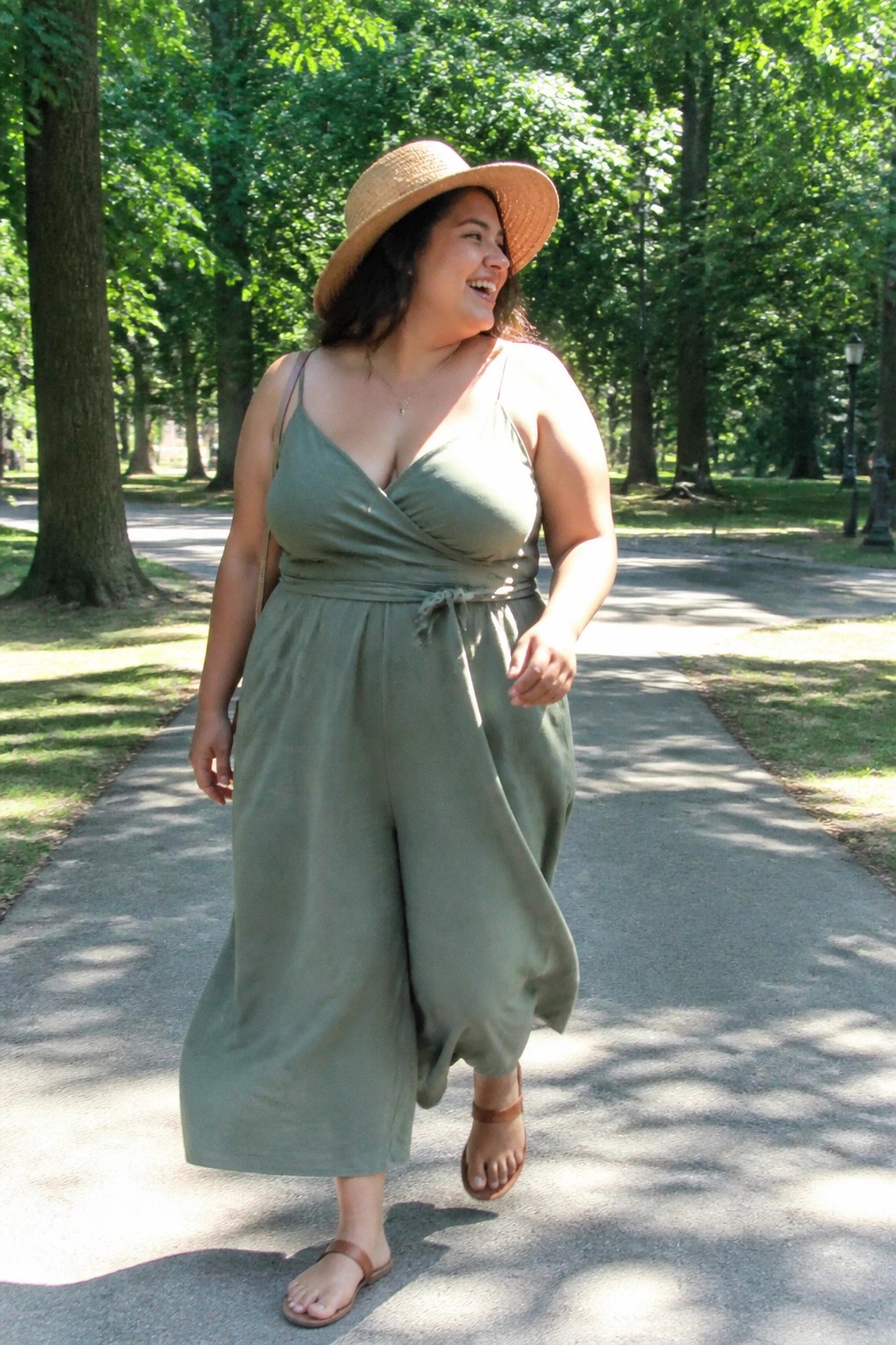 Smiling woman in sage green jumpsuit and straw hat on sunny park path.