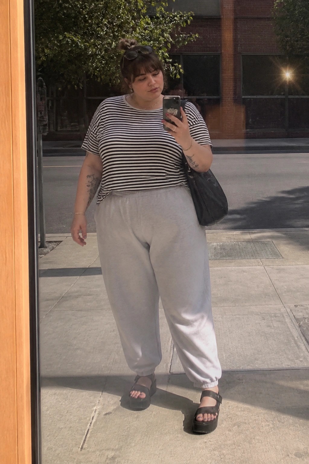 Woman with tattoos in striped shirt and gray sweatpants taking mirror selfie outside.