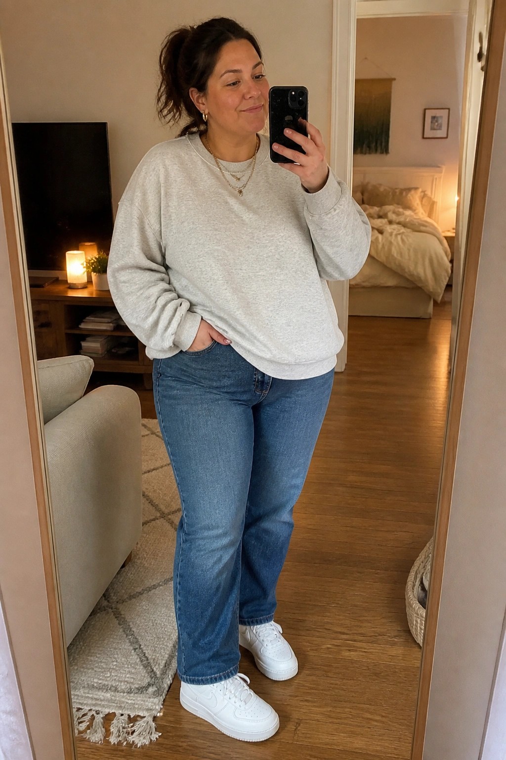 Woman in gray sweatshirt and blue jeans taking mirror selfie in home interior