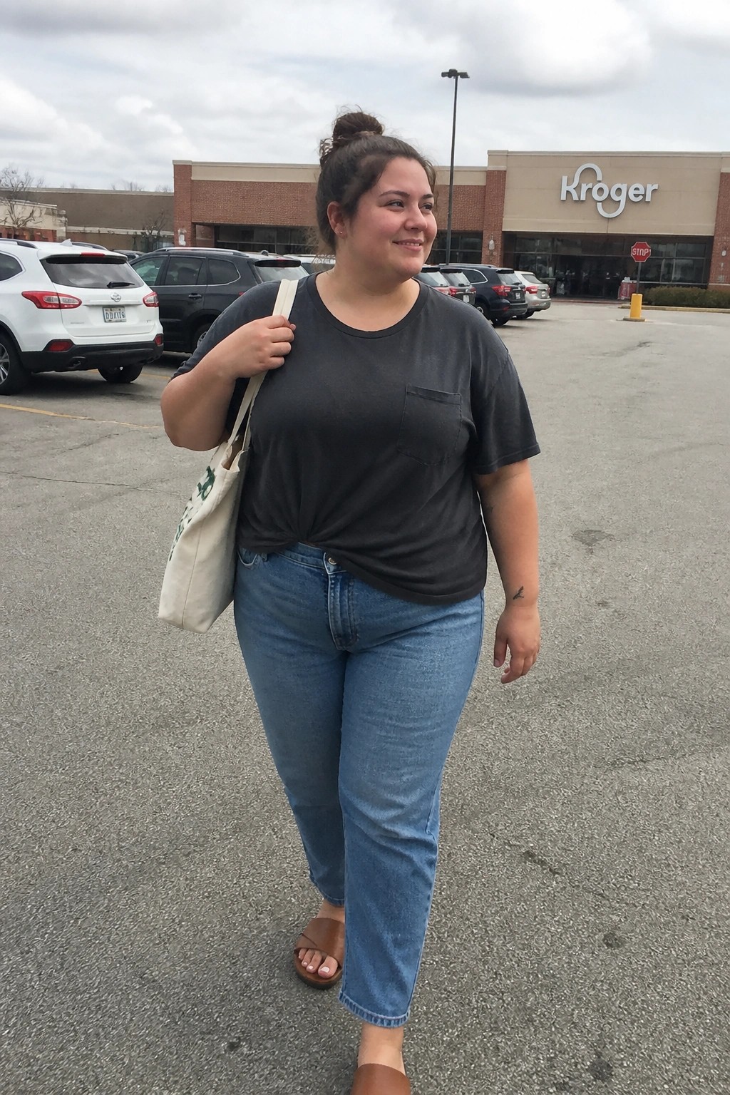 Woman carrying tote bag wearing gray t-shirt and jeans in Kroger parking lot