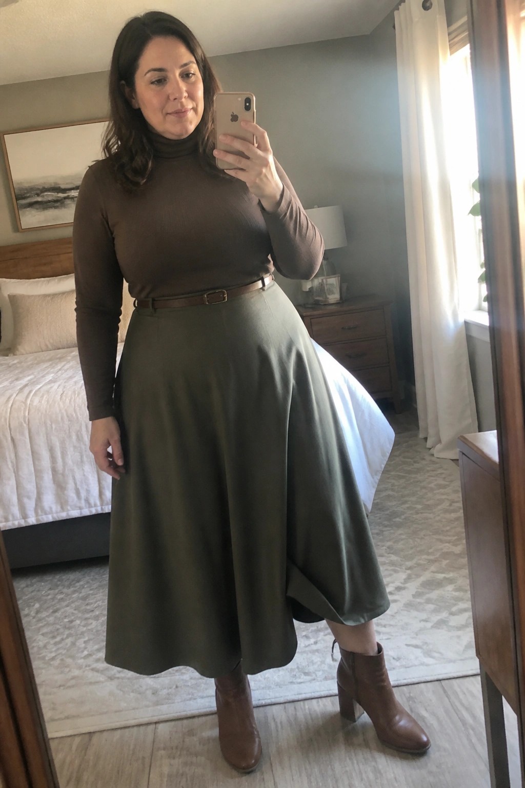 Woman wearing brown turtleneck top tucked into olive green midi skirt with brown belt and matching ankle boots while taking mirror selfie in bedroom setting