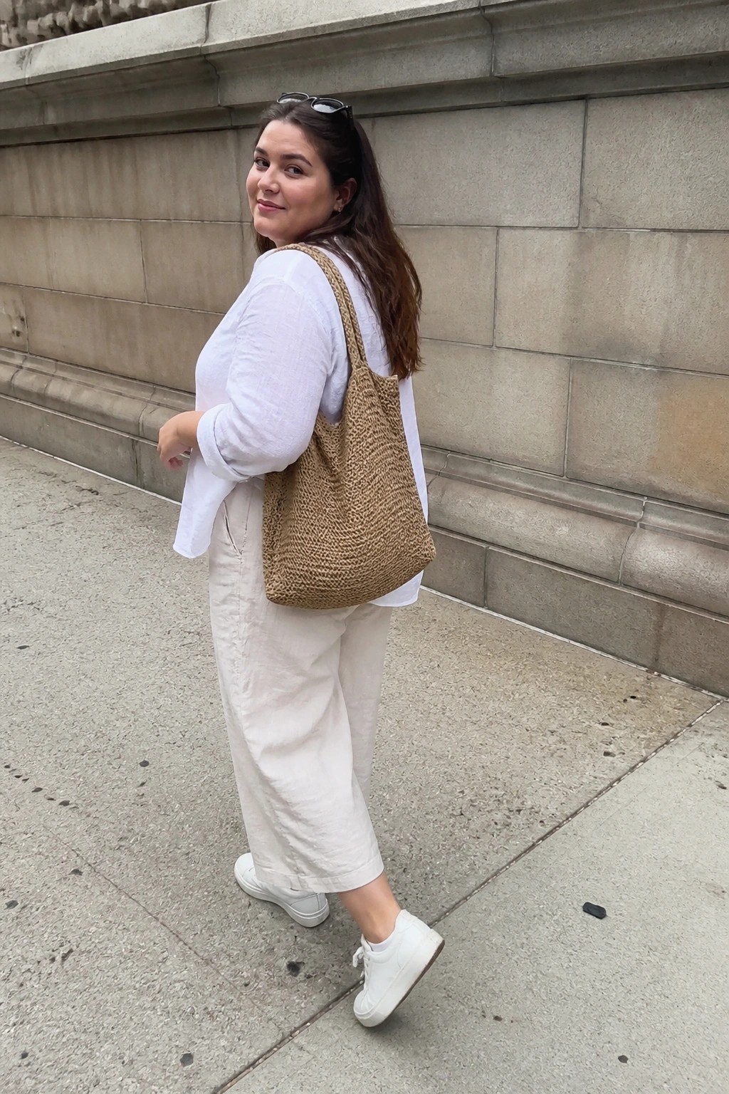 Woman in white shirt and beige pants with woven brown tote bag on sidewalk