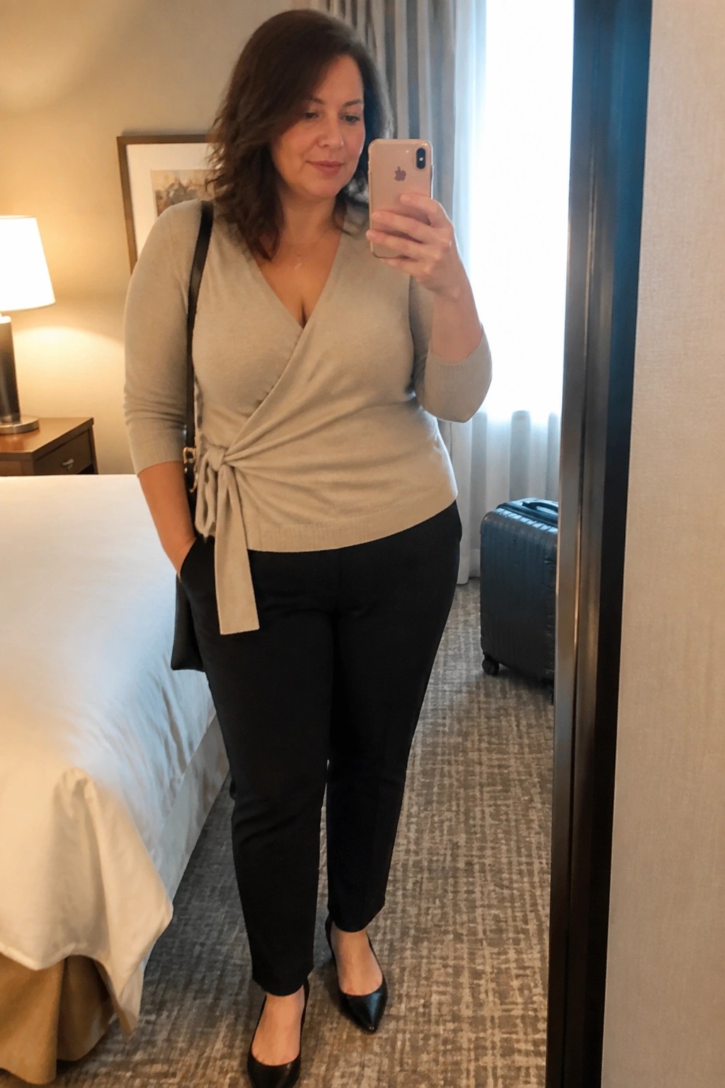 Woman wearing a beige wrap sweater tied at the waist with black pants and black heels while holding a phone for a mirror selfie.