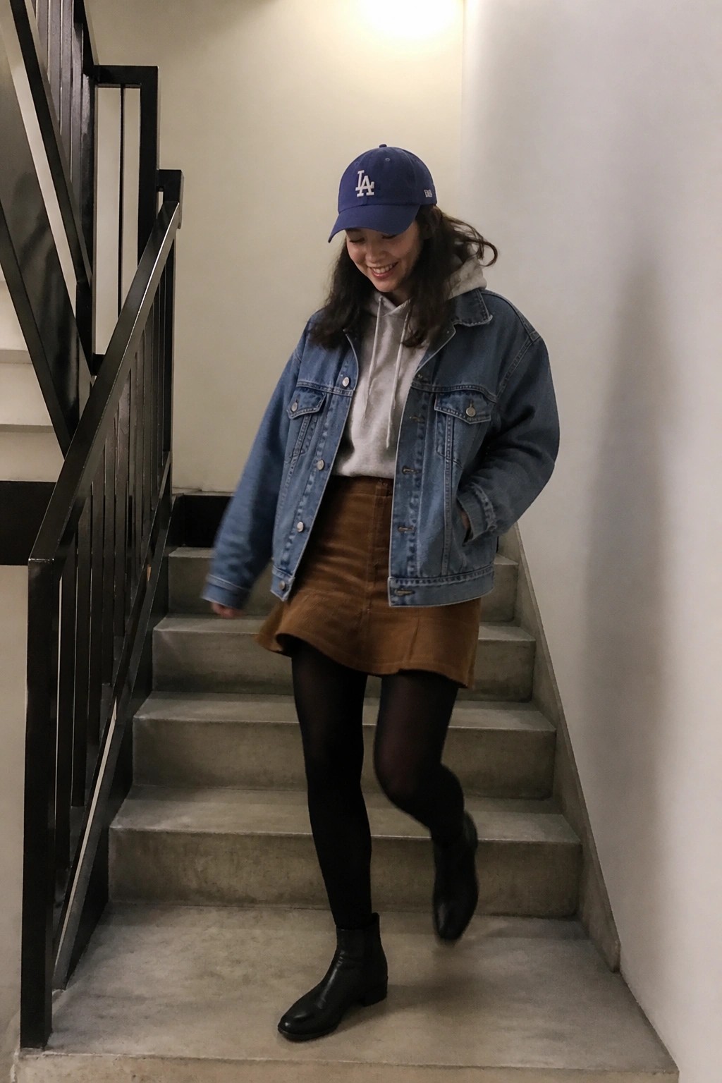 Woman in blue cap and denim jacket smiles on stairs wearing brown skirt