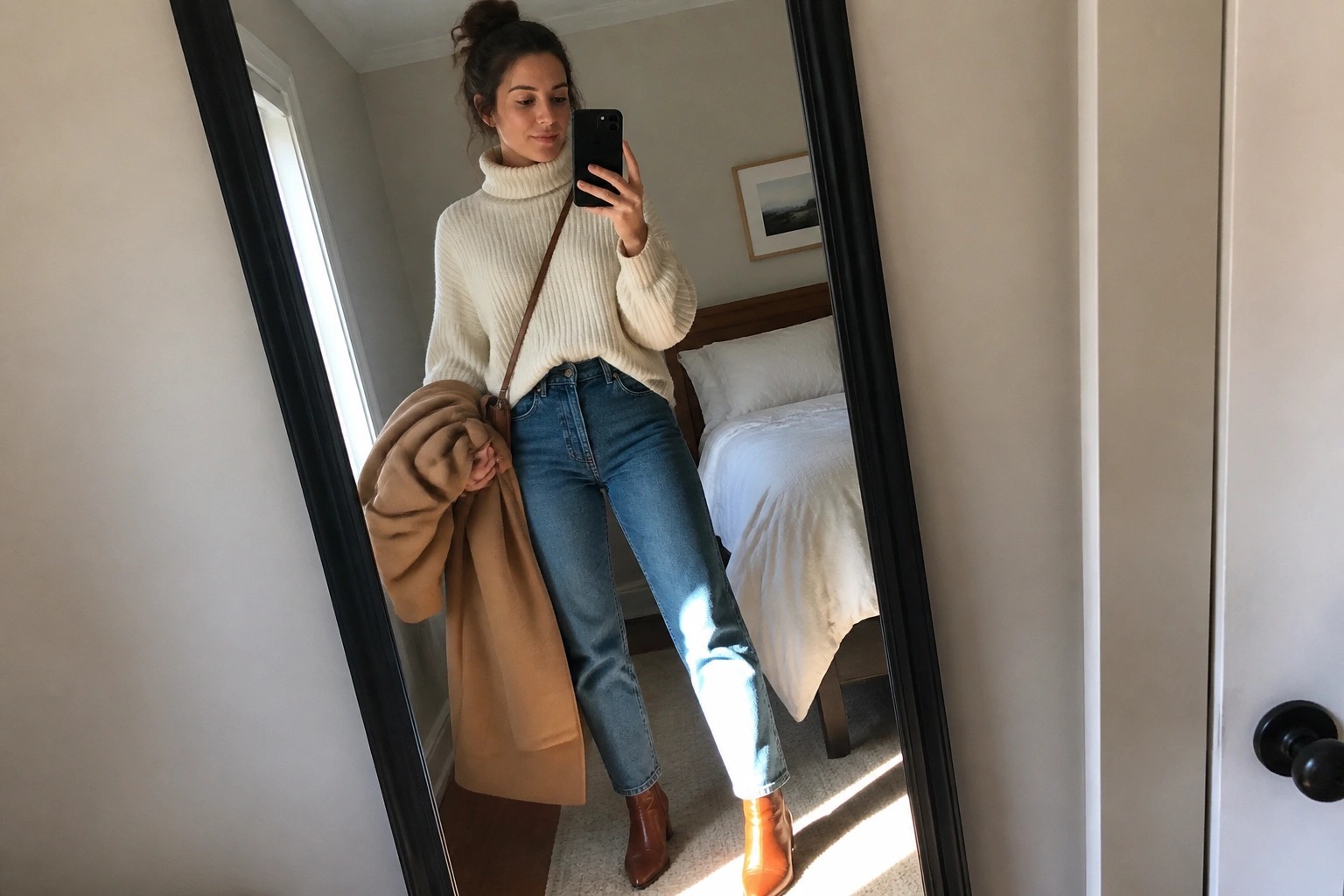 Woman in cream turtleneck sweater and jeans taking bedroom mirror selfie