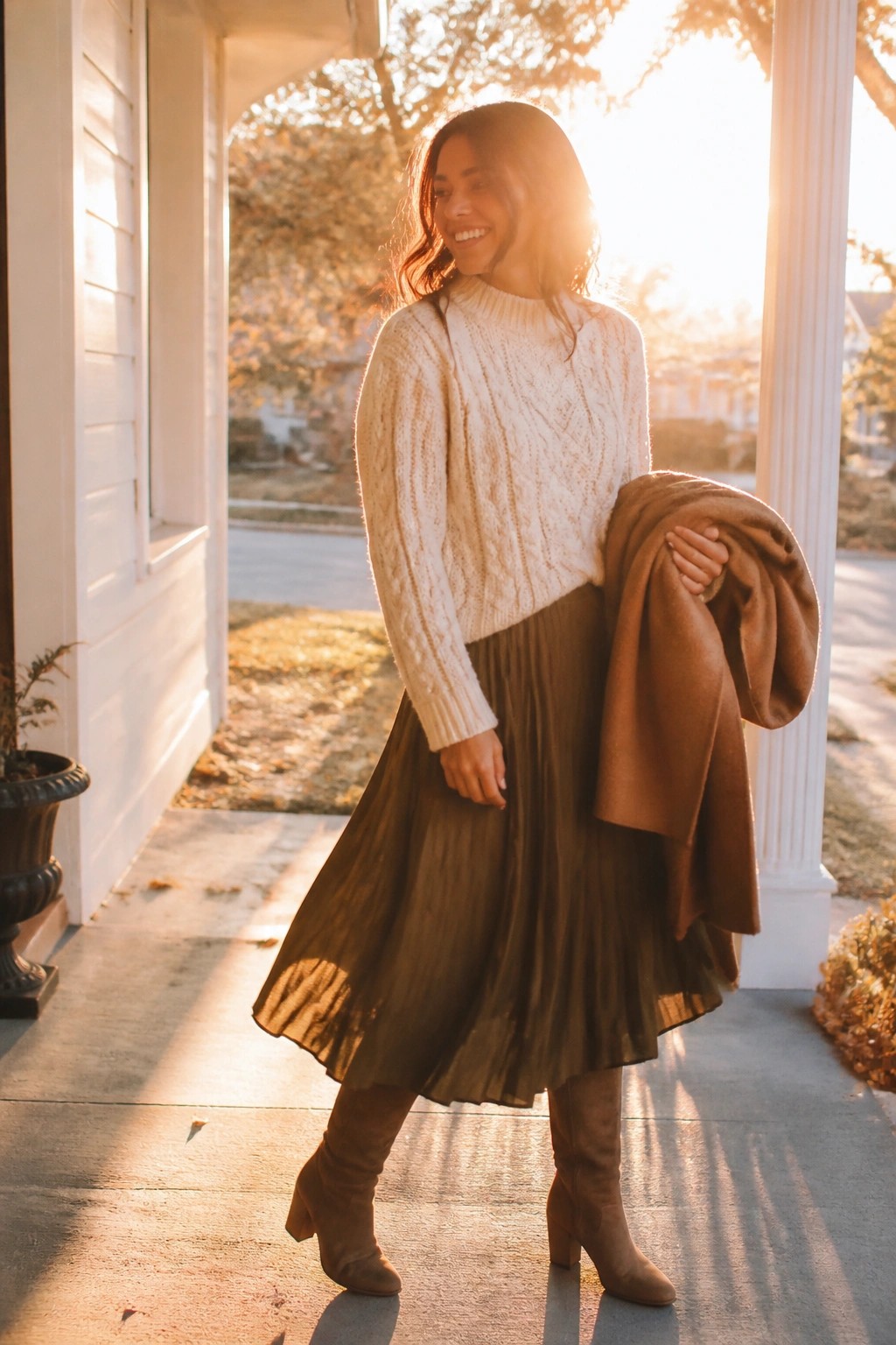 Woman wearing cream cable knit sweater tucked loosely into olive green pleated midi skirt with brown suede knee high boots and holding a brown coat over one arm.