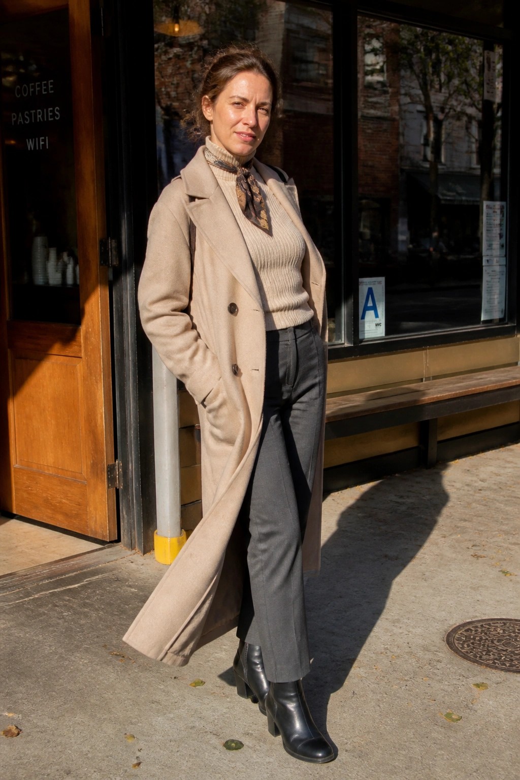 Woman wearing a long beige coat open over a beige turtleneck sweater with a patterned scarf, gray trousers, and black ankle boots.