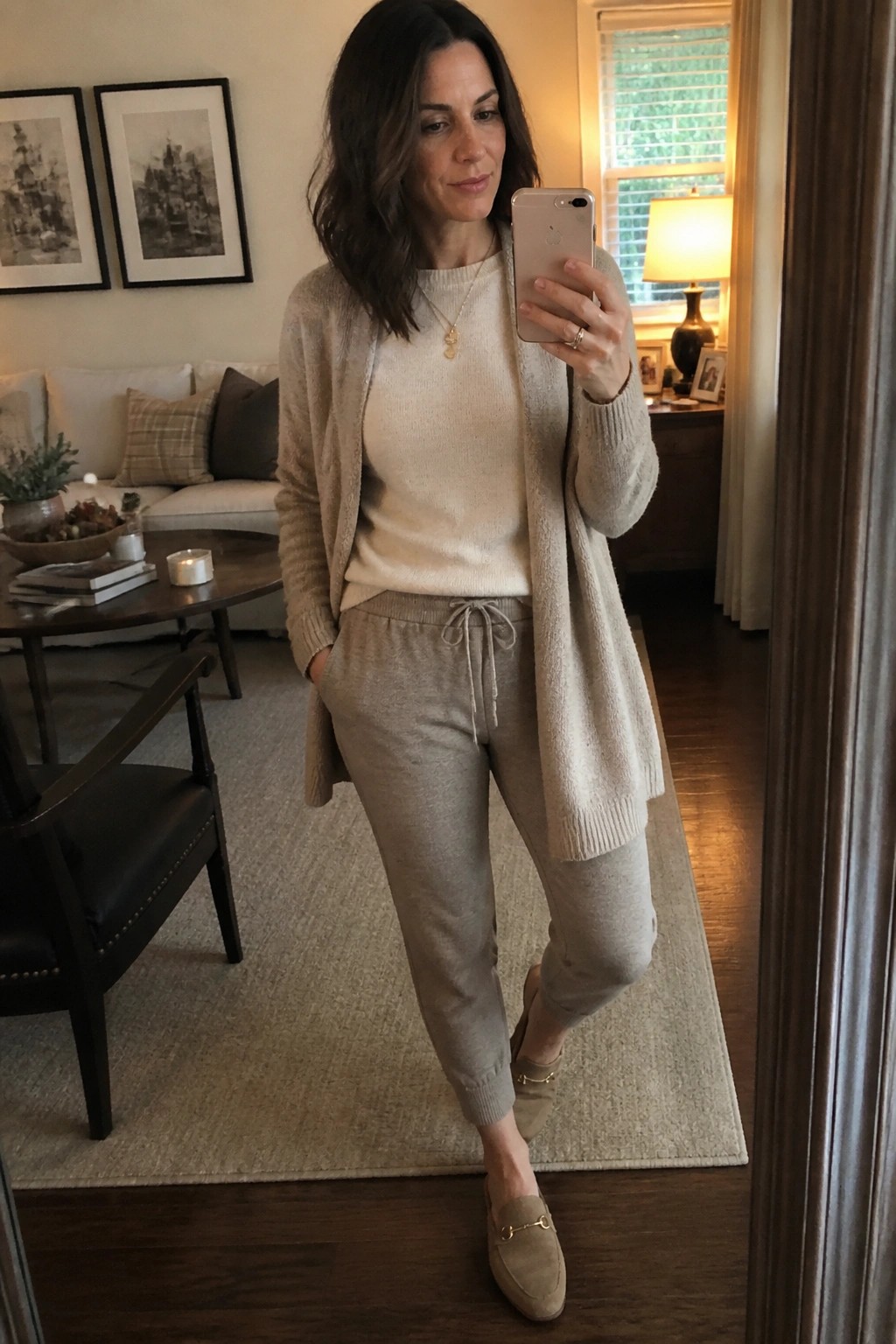 Woman in beige cardigan and joggers taking mirror selfie in living room
