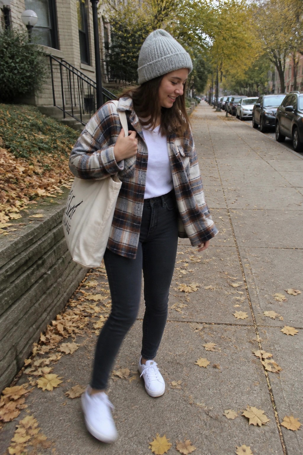 Woman in plaid jacket and gray beanie walks on autumn sidewalk with tote bag