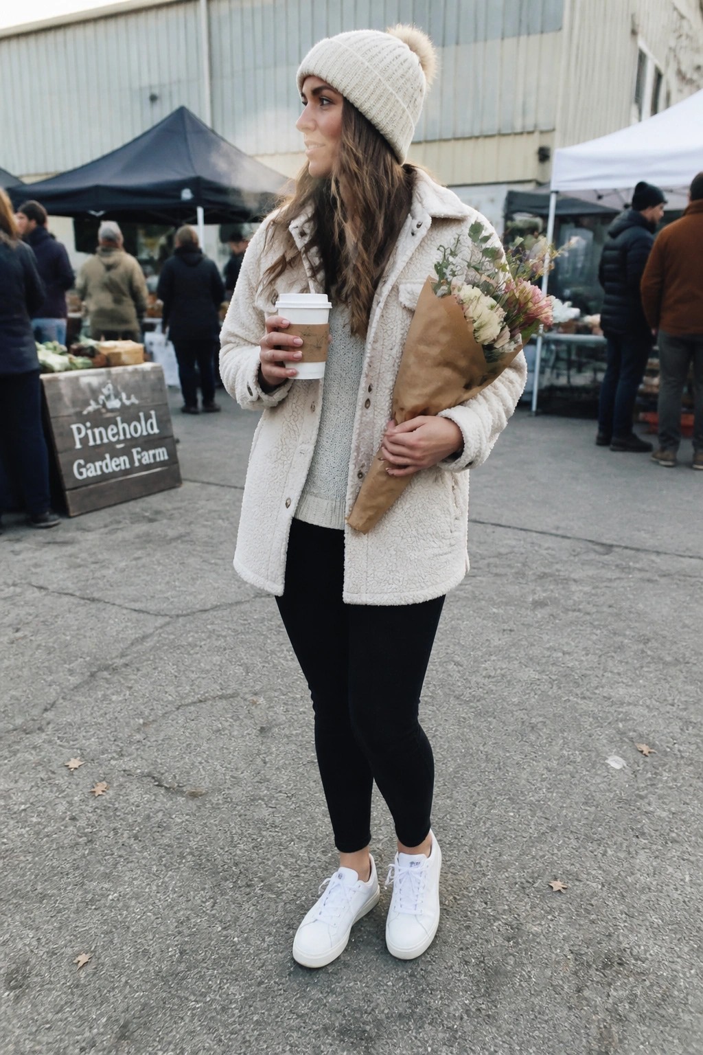 Woman wearing cream sherpa jacket layered over gray knit sweater with black leggings and white sneakers holding coffee and flowers.