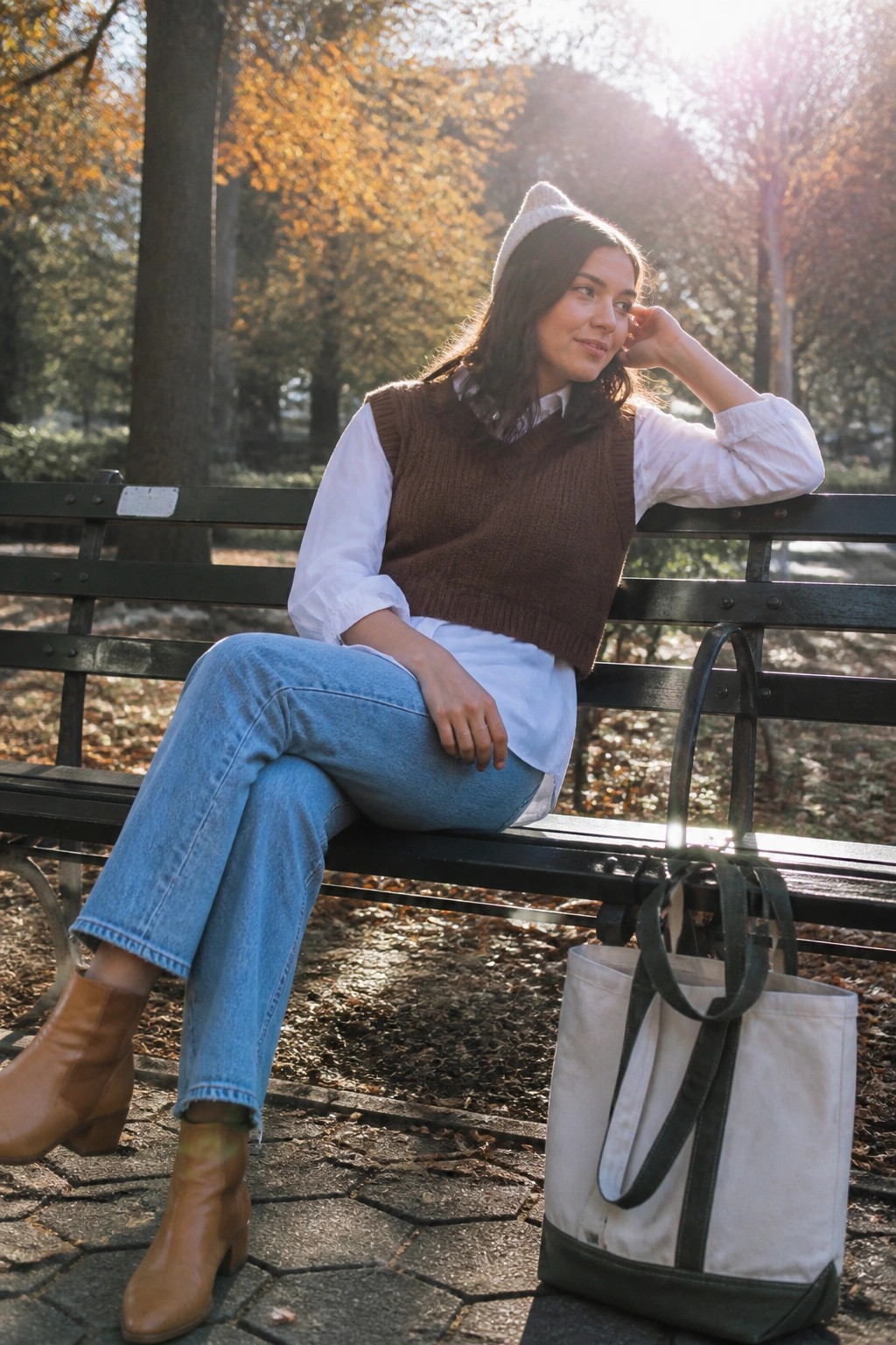 Woman in brown sweater vest and white beanie sits on park bench with tote bag