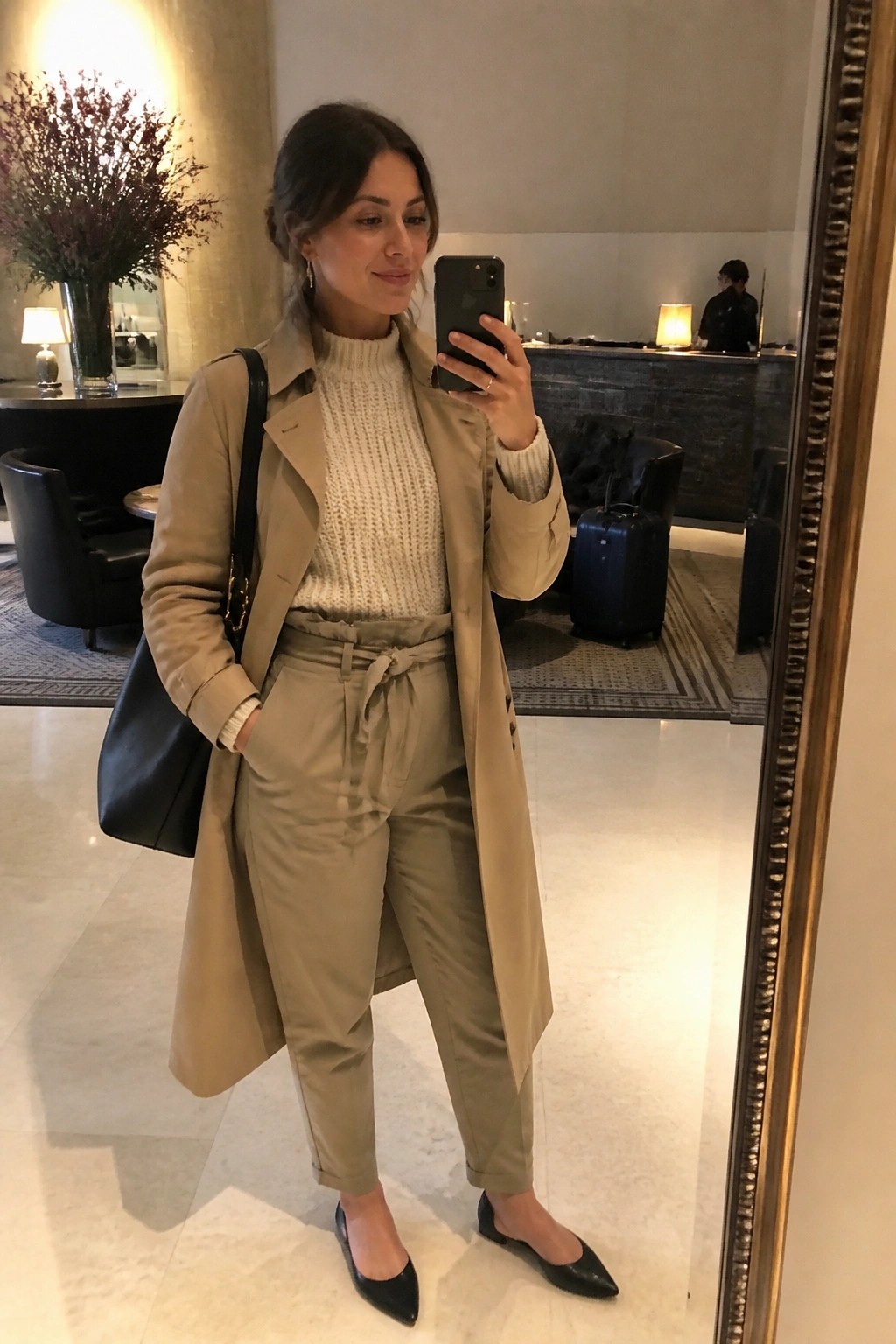 Woman in beige trench coat and white sweater taking mirror selfie in lobby