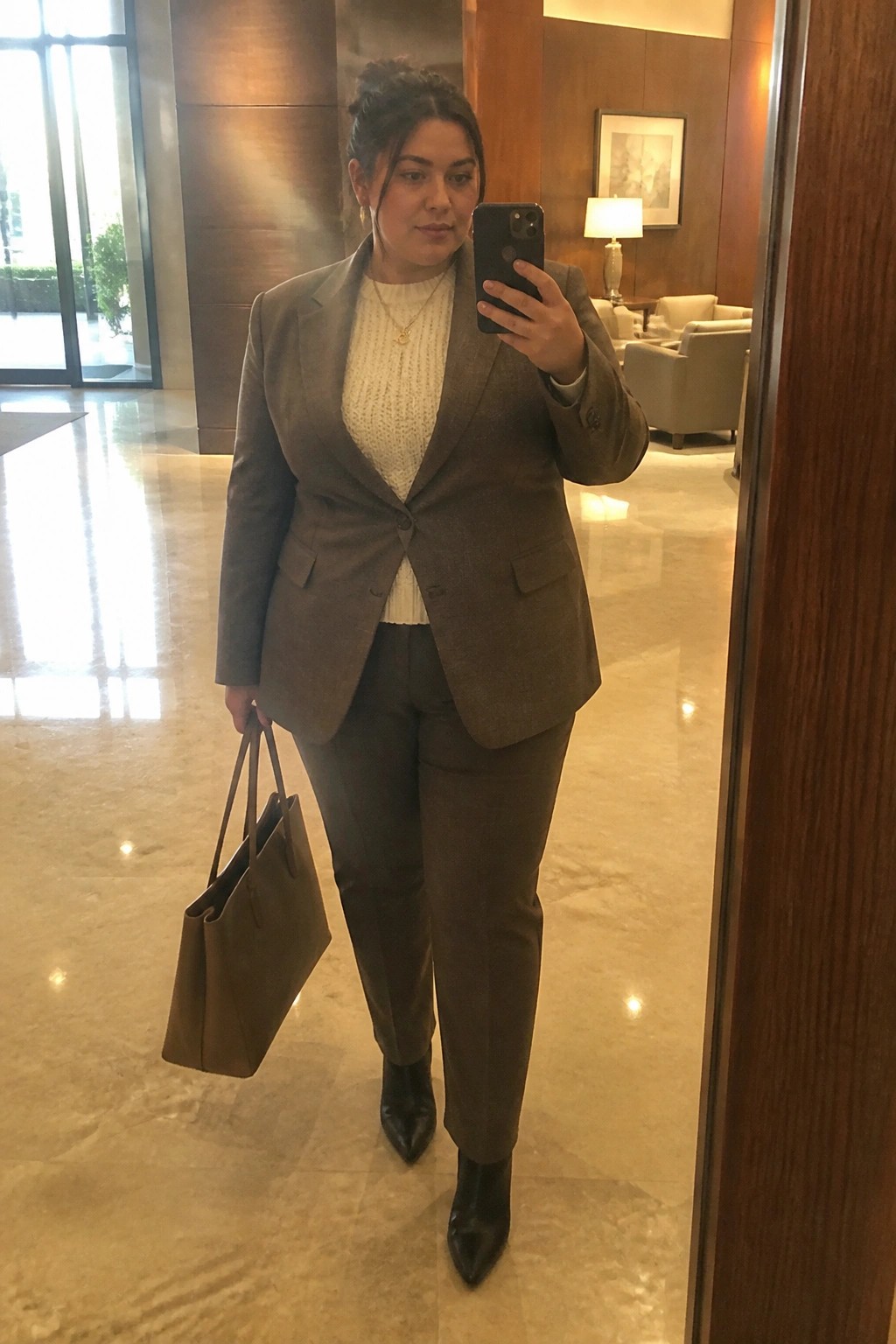 Woman in brown suit taking mirror selfie in marble lobby holding phone and bag.