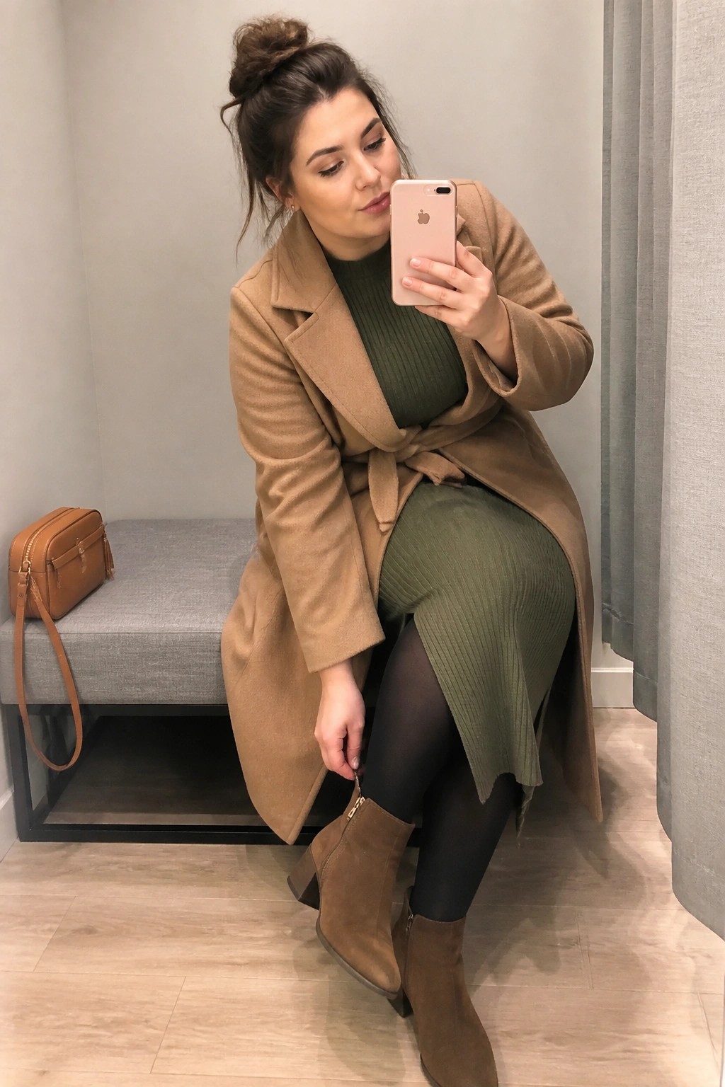 Woman in tan coat over green dress taking selfie on bench with iPhone