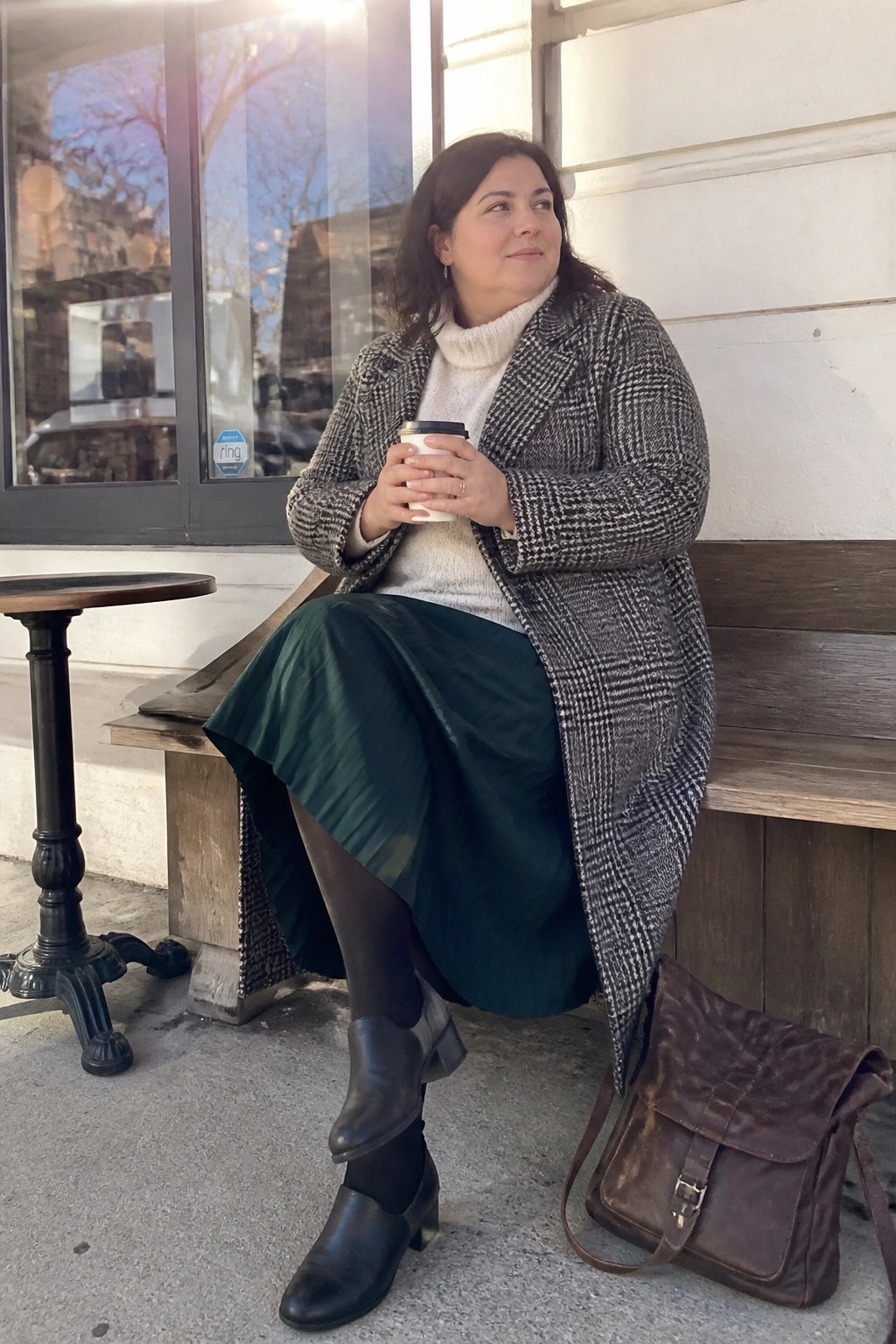 Woman in houndstooth coat and green skirt holds coffee on wooden bench.