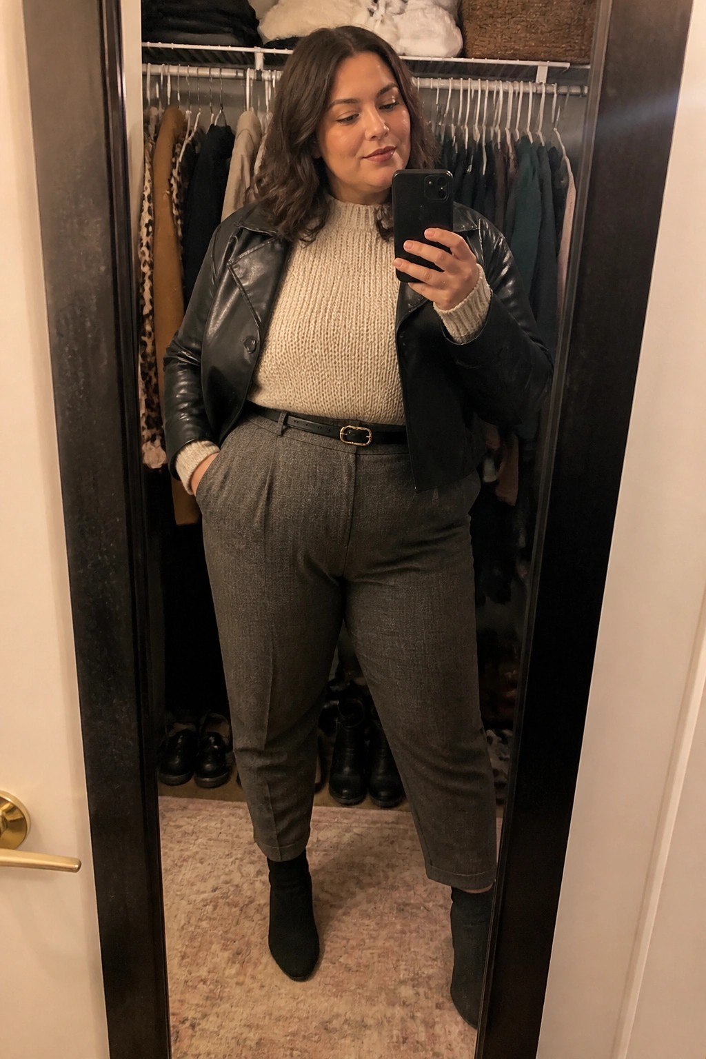 Woman in beige sweater and black leather jacket taking mirror selfie in closet