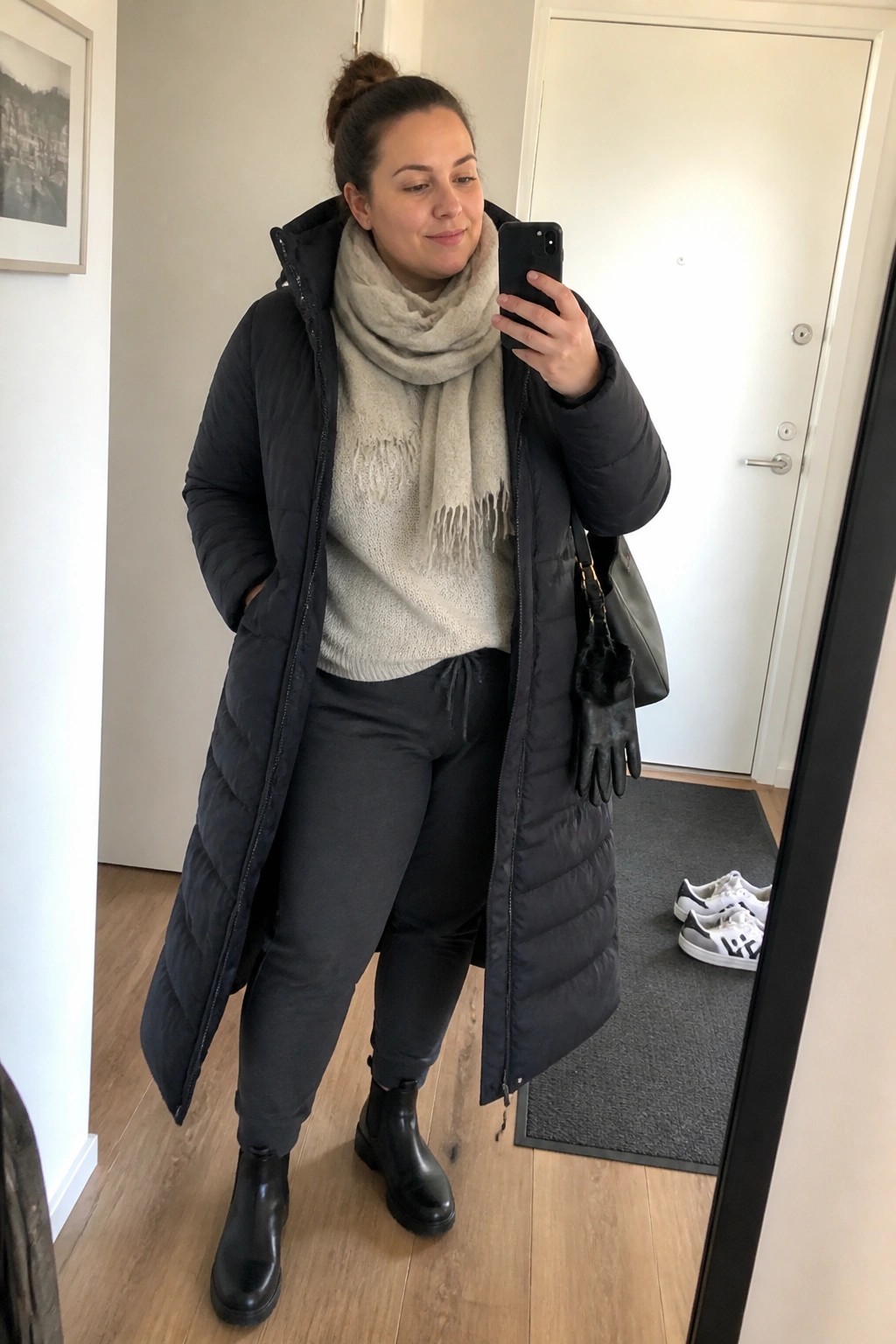 Woman in long black puffer coat and scarf taking mirror selfie.