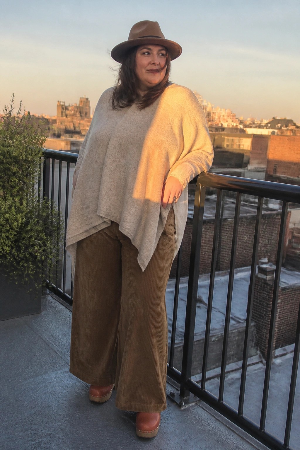 Woman in beige poncho, brown hat, and corduroy pants on city balcony at sunset