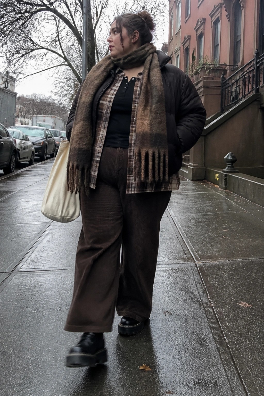 Woman in plaid shirt, fuzzy scarf, and corduroy pants on wet city sidewalk