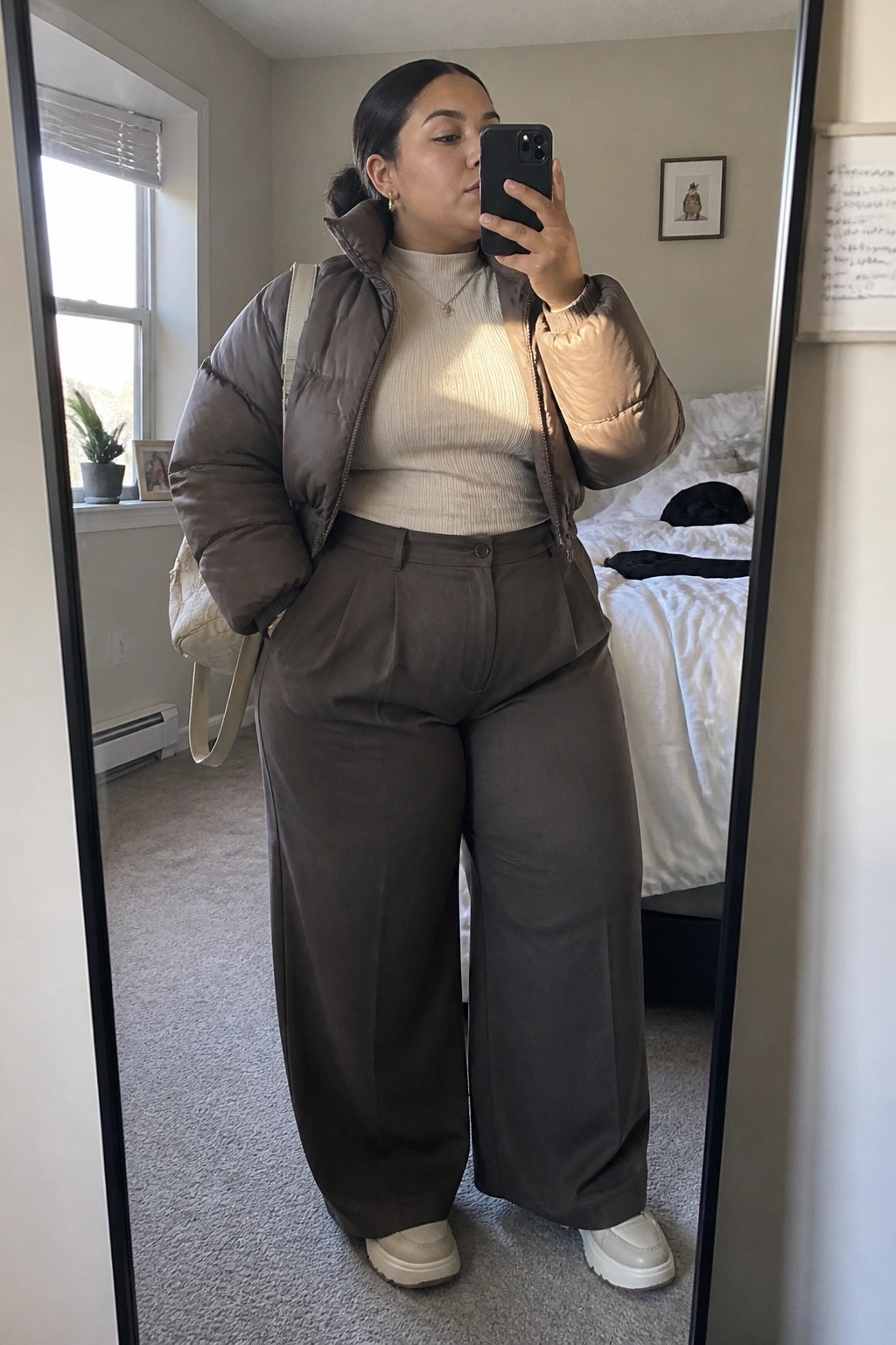 Woman in brown puffer jacket, beige sweater, and wide trousers taking mirror selfie
