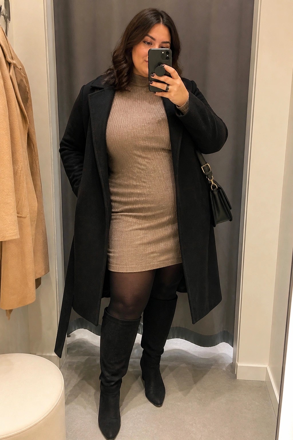 Woman in black coat over beige knit dress taking mirror selfie in fitting room