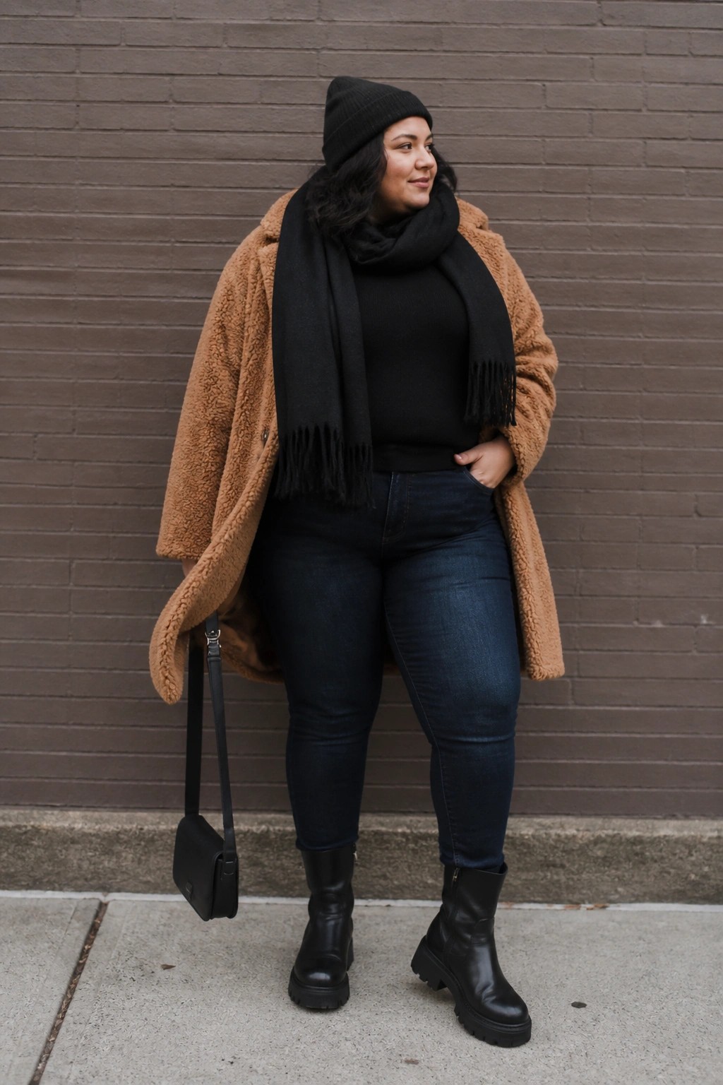 Plus size woman wearing an open brown teddy coat over a black turtleneck sweater and fringed black scarf with dark blue jeans and black combat boots, holding a small black crossbody bag.